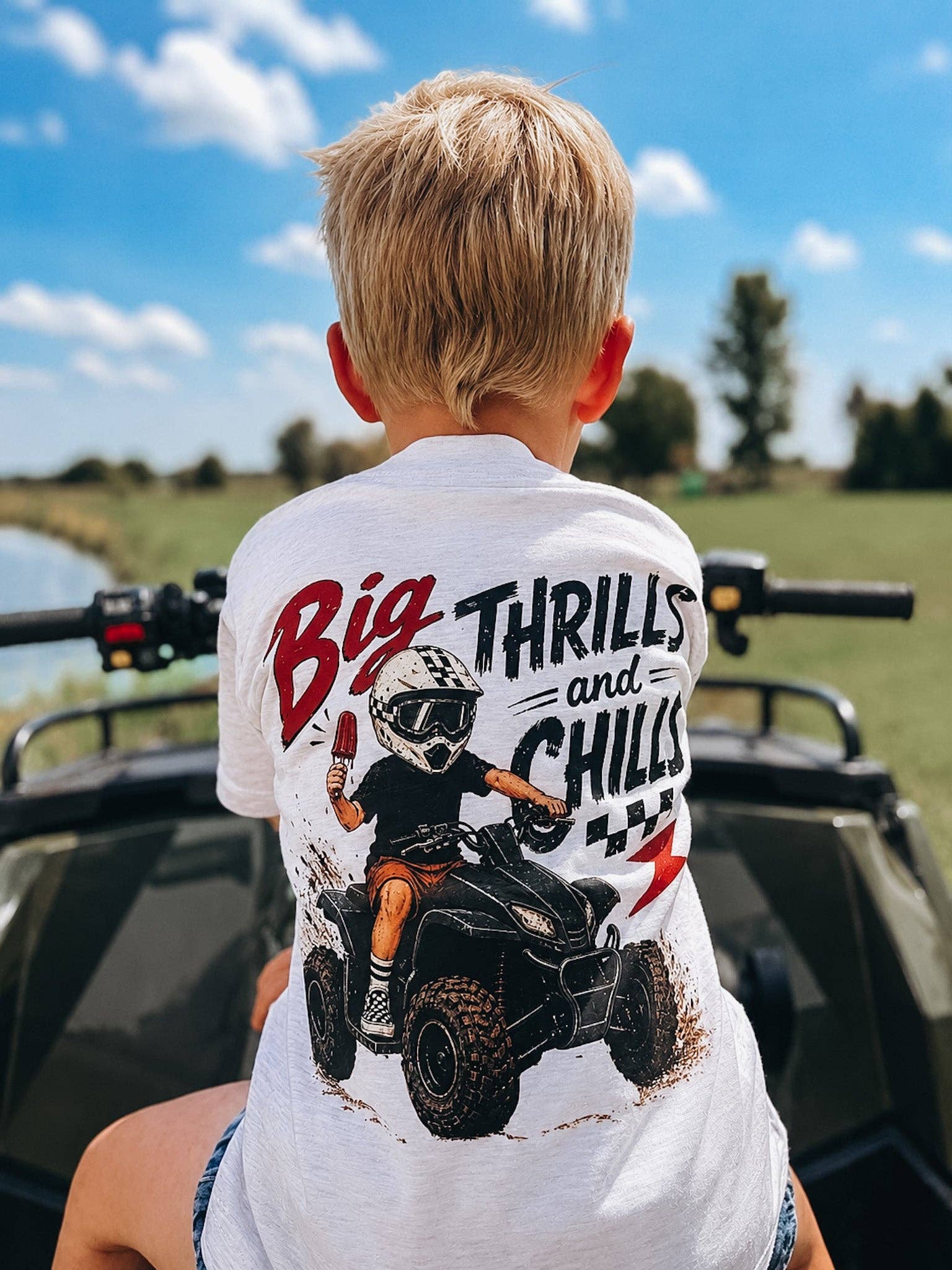 A young boy with blond hair sits on an ATV outdoors on a sunny day, wearing the Raising Brave Big Thrills and Chills Toddler Tee, which features a cartoon rider and the text "Big Thrills and Chills" on the back. Green grass and a blue sky complete the scene.