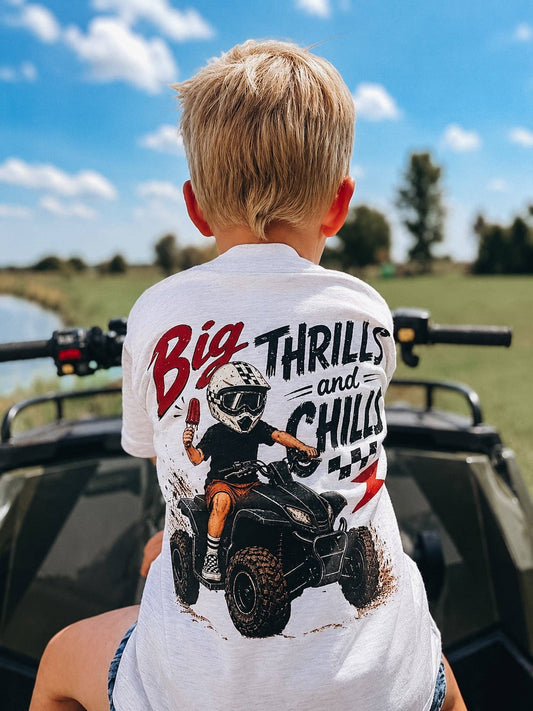 A young boy with blond hair sits on an ATV outdoors on a sunny day, wearing the Raising Brave Big Thrills and Chills Toddler Tee, which features a cartoon rider and the text "Big Thrills and Chills" on the back. Green grass and a blue sky complete the scene.