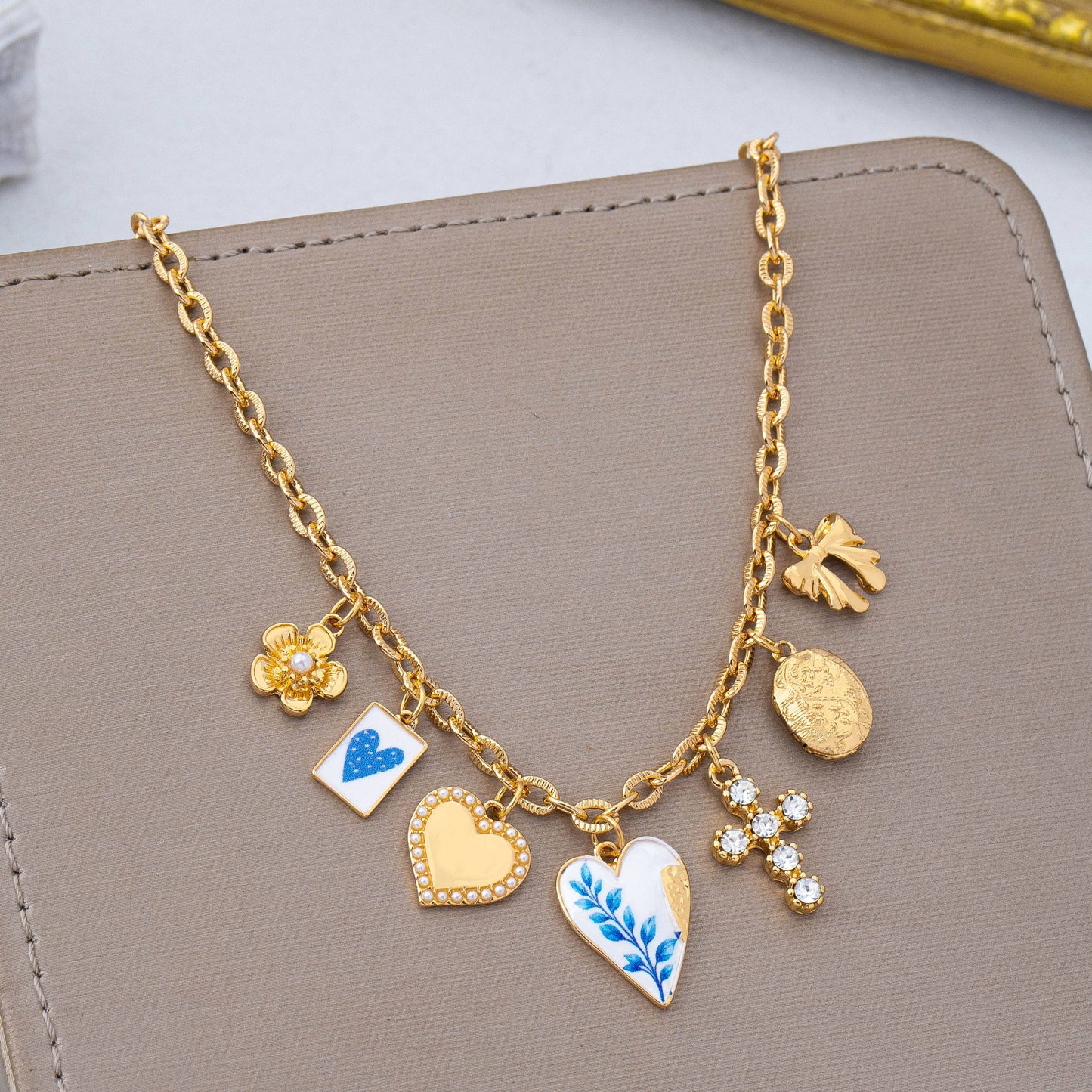The Blue Vintage Coquette Charm Necklace by JOSSLYN by wall to wall features a gold chain adorned with six unique charms—including a flower, a card with a blue heart, a gold heart, a white heart detailed with a blue leaf design, a cluster of daisies, and an oval medallion—beautifully displayed on a taupe surface.