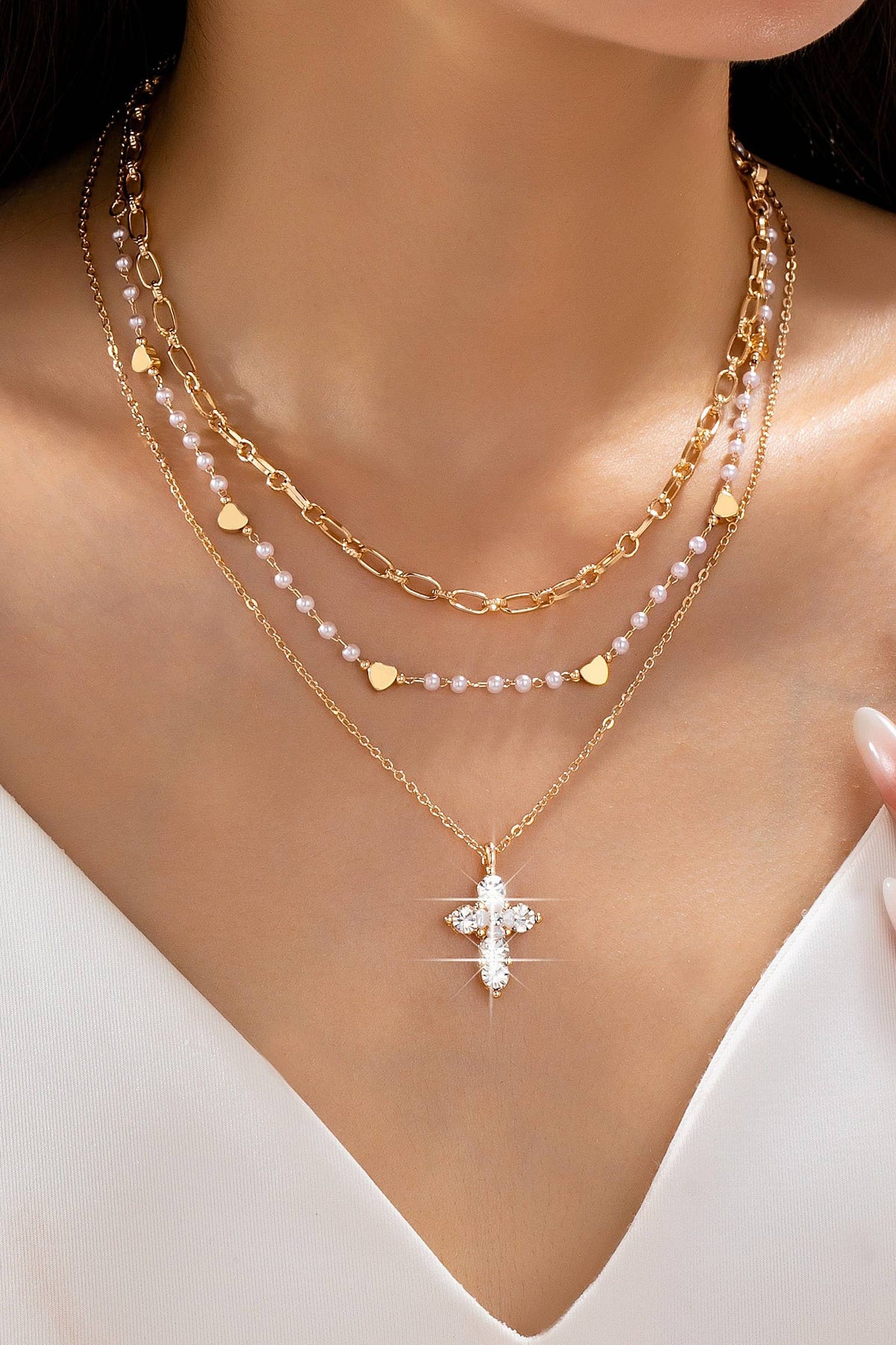 A woman wearing the JOSSLYN by wall to wall Multi-Strand Pearl & Crystal Cross Necklace, featuring layered strands with heart charms, beads, and a sparkling cross pendant, over a white top.