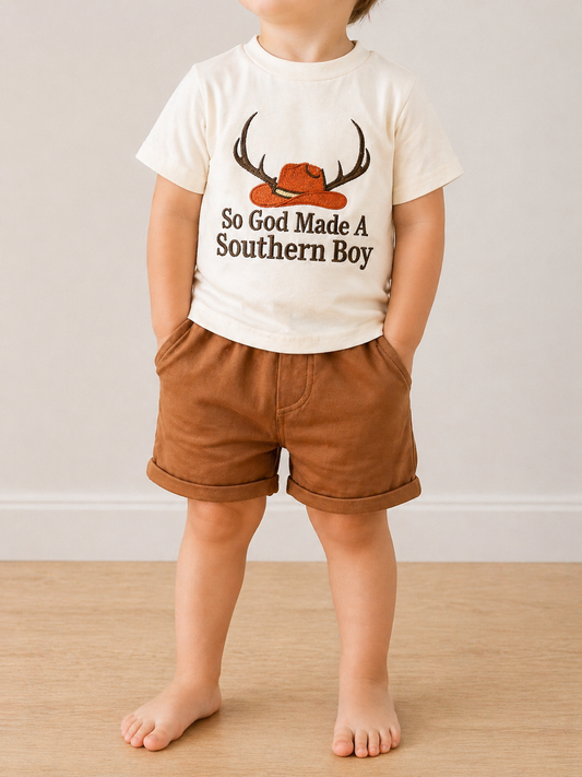 A young child stands barefoot on a wood floor, hands in pockets, wearing the Rustic Roots Short Set by Tiny Tot Bundles: brown shorts and a white tee featuring a hat-and-antler graphic with "So God Made A Southern Boy" text.