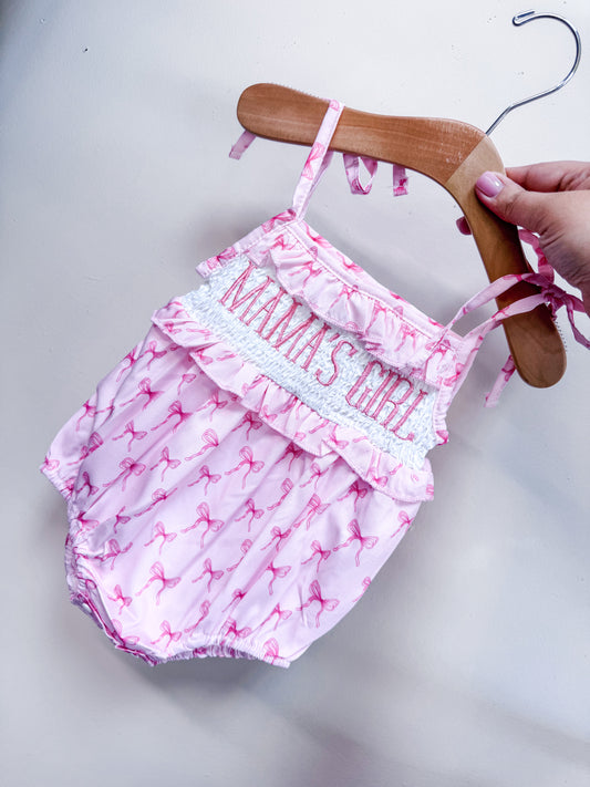 The Mama's Coquette Cutie Romper by Tiny Tot Bundles features pink fabric, a flamingo pattern, ruffles, and “MAMAS GIRL” embroidery. Shown on a wooden hanger held by a hand with pink nail polish.