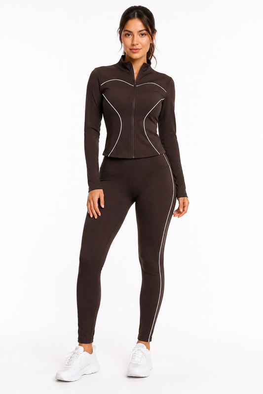 A woman stands in the Color 5 Chocolate Brown Active Set— a fitted athletic tracksuit featuring white piping, a zip-up jacket, and matching leggings. She pairs the outfit with white sneakers and smiles slightly against a plain white background.