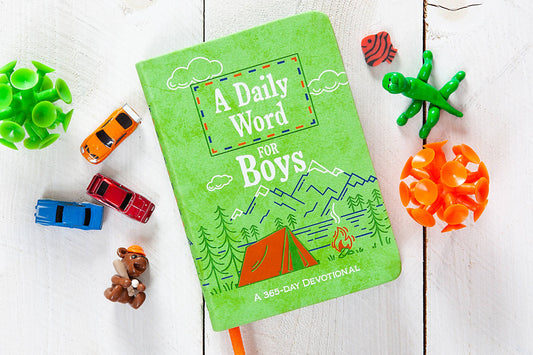 A green “A Daily Word for Boys Book” from BroadStreet Publishing Group, LLC rests on a white wooden surface, surrounded by toy cars, animal figures, a green figure, and bright orange game pieces.