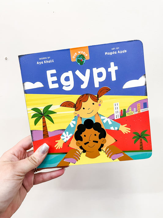 A hand holds Barefoot Books’ "Our World: Egypt," a colorful children’s book featuring a smiling girl on a boy’s shoulders, with palm trees and buildings in the background. The book is by Aya Khalil and Magda Azab.