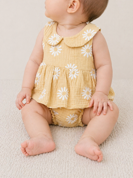 A baby sits on a light carpet, wearing the Tiny Tot Bundles Sunny Bloom Romper—a yellow sleeveless outfit with white daisy print and a peter pan collar. The baby is facing slightly sideways, hands resting on the floor.