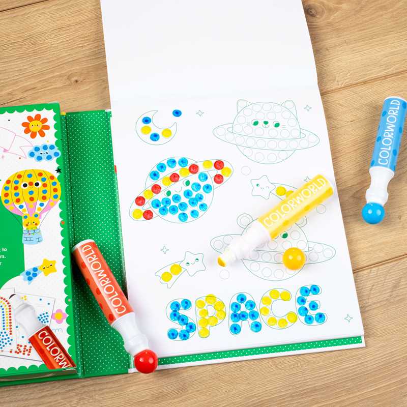 A coloring activity with ColorWorld: Dot Art Fun Activity Book by Simon & Schuster is in progress, featuring pages with planets, stars, and “SPACE” in dots. Three COLORWORLD dot markers—red, blue, and yellow—lie nearby on a wooden surface.