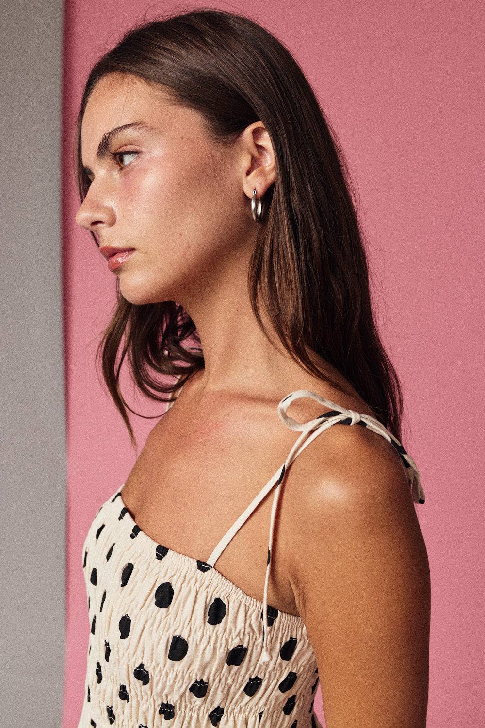 A woman with long brown hair, wearing the Papermoon Polka Dot Smocked Mini Dress, stands in front of a pink and gray background, looking to the left.
