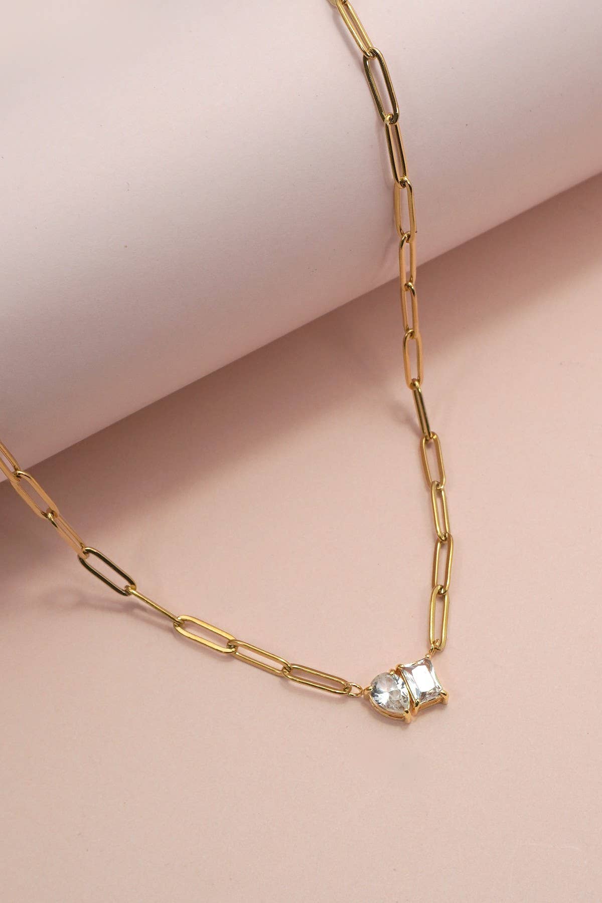 A Gold Rhin Necklace by JOSSLYN by wall to wall, featuring a clear gemstone pendant on a gold paperclip chain, rests on a light pink surface next to a rolled piece of paper.