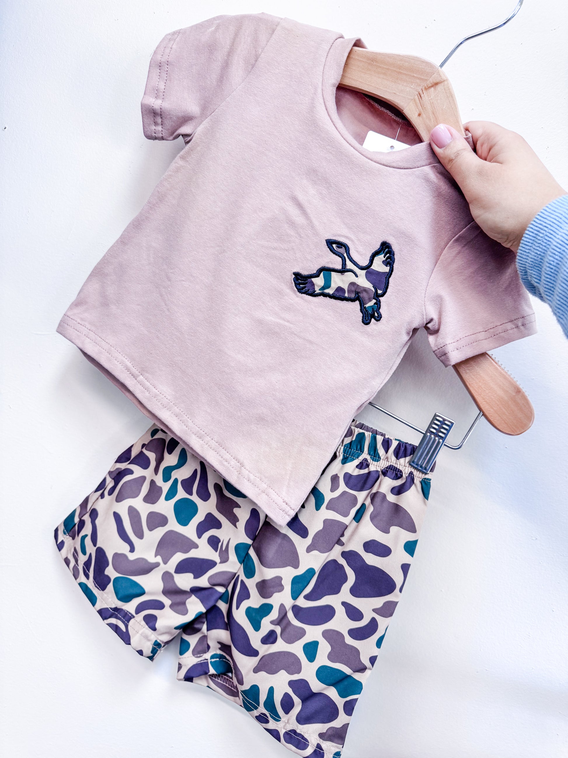 A hand displays the Little Hunt Duo Boys Set by Tiny Tot Bundles—a light pink short-sleeve shirt with a camouflage bunny patch, paired with matching purple, green, and beige camouflage shorts.