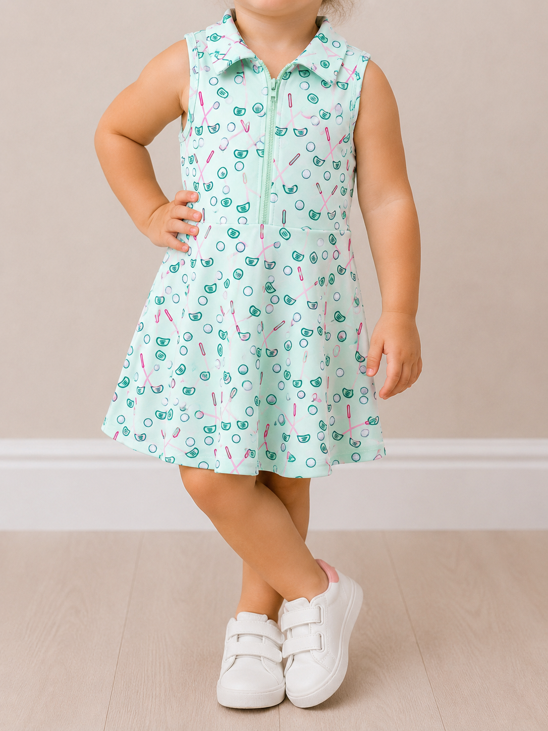 A young child models the Tiny Tot Bundles Green Quarter Zip Golf Dress, standing with legs crossed and one hand on hip. Paired with white sneakers, the face is not shown against a neutral background.
