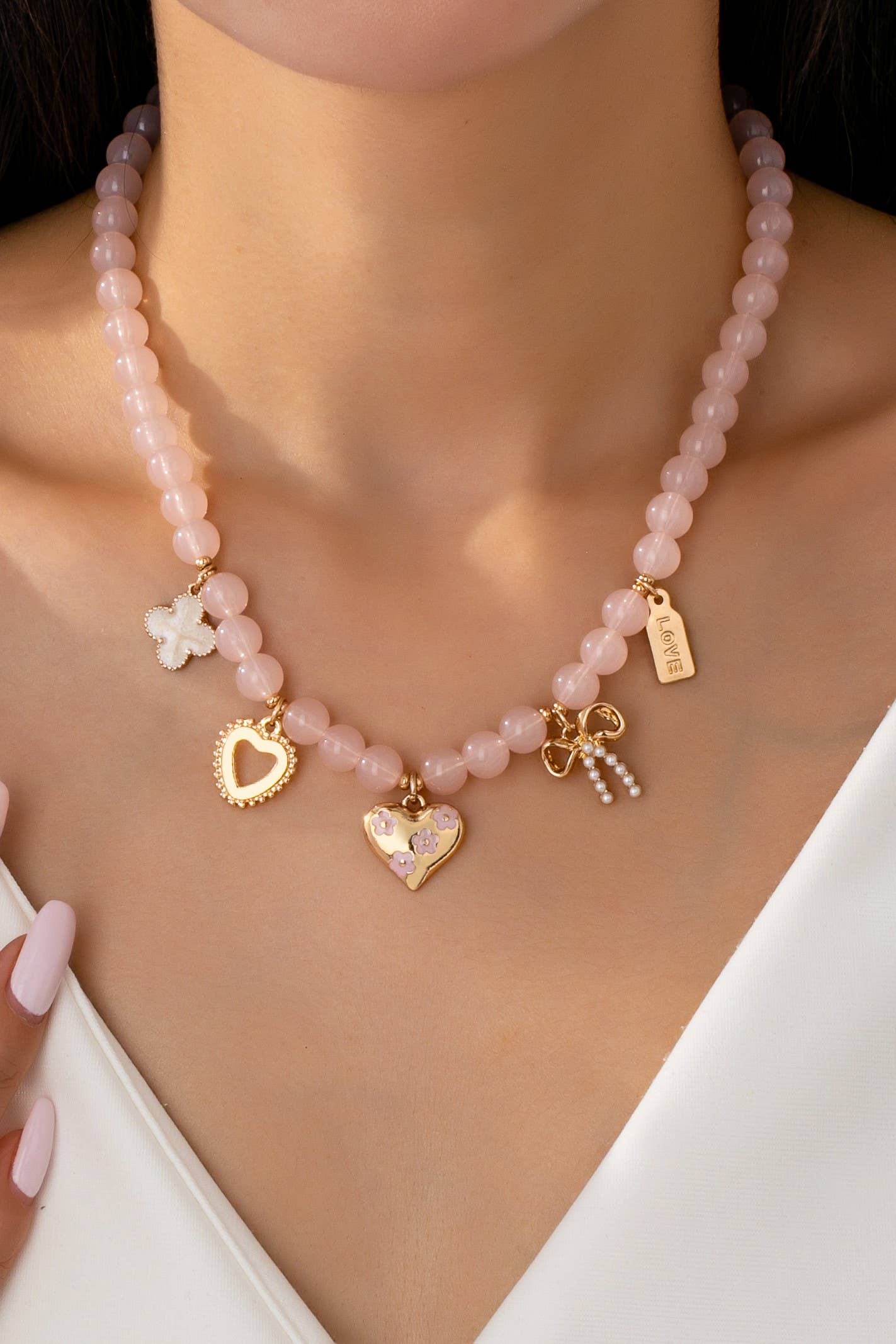 A woman wearing a white top showcases the JOSSLYN by wall to wall Beaded Charm Necklace, featuring pink beads and five gold and pink charms—a heart, a bow, a clover, another heart, and a “love” tag. One hand with pale pink nails rests near the necklace.