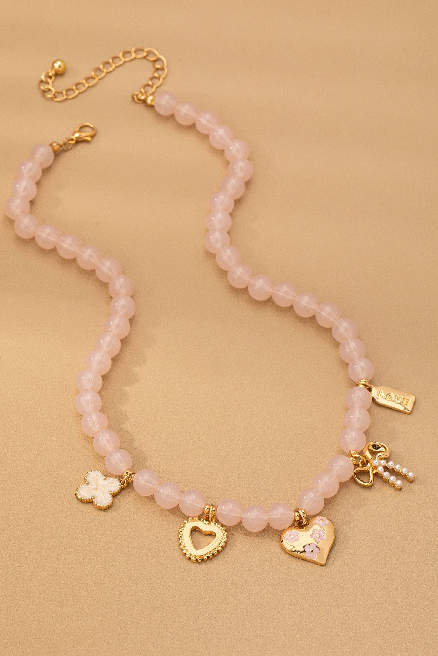 The Beaded Charm Necklace by JOSSLYN by wall to wall features pink beads and five gold charms—a clover, a heart outline, a heart adorned with stones, a tag engraved with LOVE, and a clustered charm with a key, gold heart, and pearls—beautifully displayed on a tan surface.