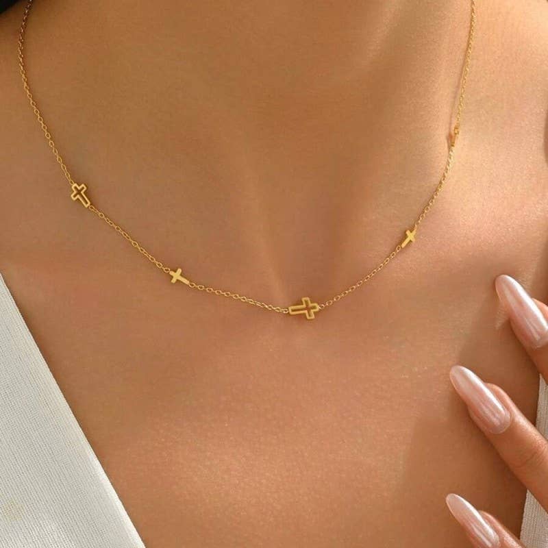 A close-up of a person wearing the JOSSLYN by wall to wall Dainty Cross Necklace, featuring small gold cross charms, paired with a white top. Their nails are manicured and glossy, with one hand gently touching their collarbone.