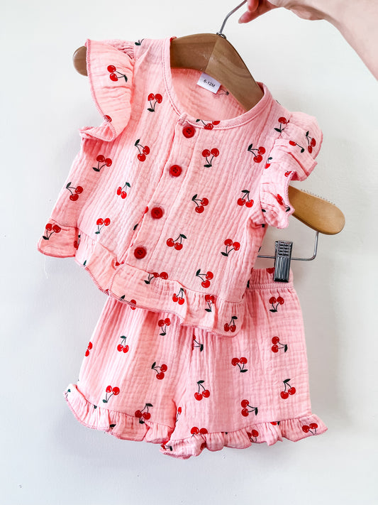The Linen Chery Pink Set by Tiny Tot Bundles, featuring a pink ruffled cherry-print top and matching shorts, is displayed on a wooden hanger against a white background.