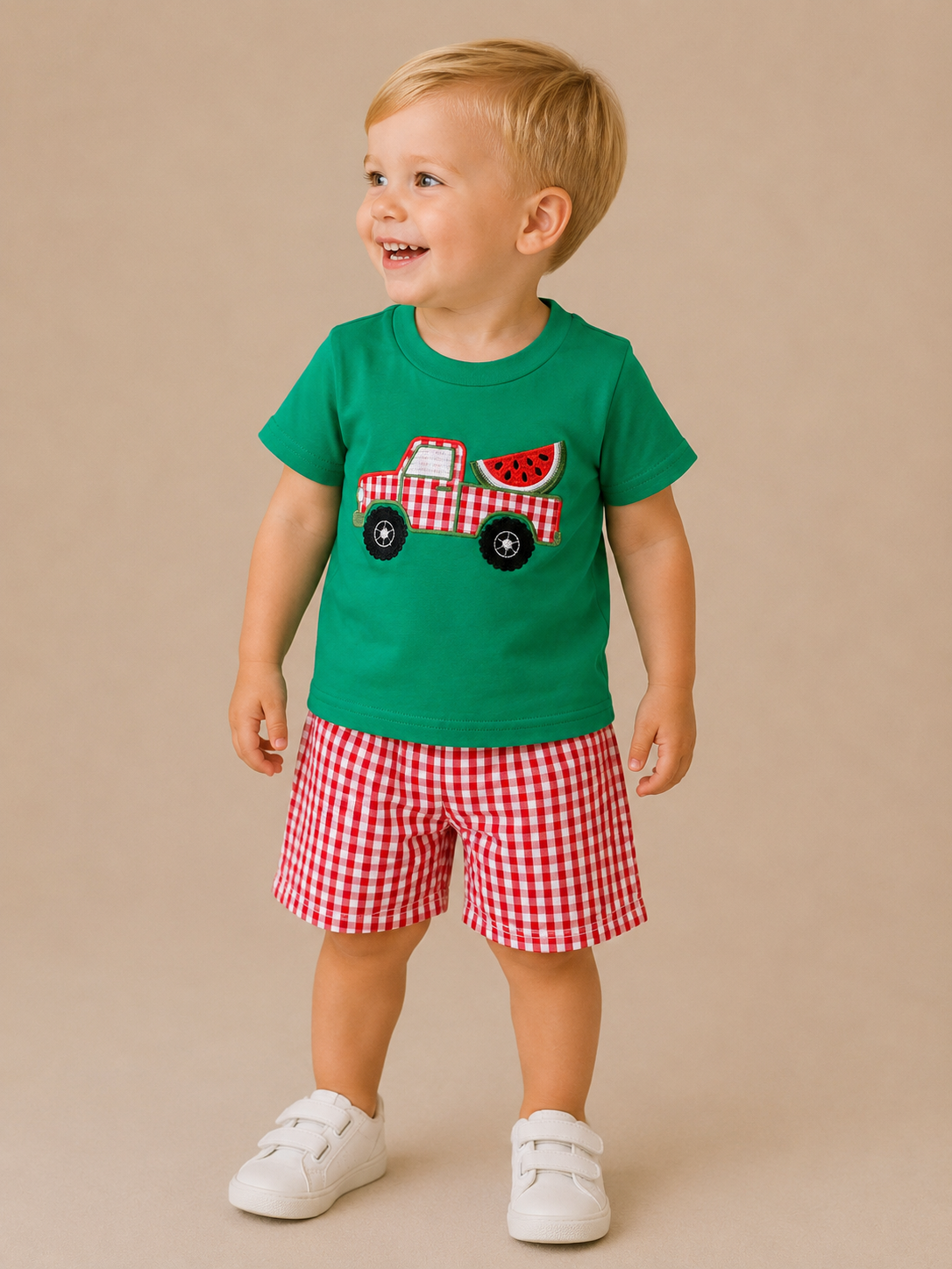 A smiling toddler models the Tiny Tot Bundles Watermelon Wagon Set, featuring a green tee with a red truck and watermelon graphic, and red checkered shorts, set against a beige background.