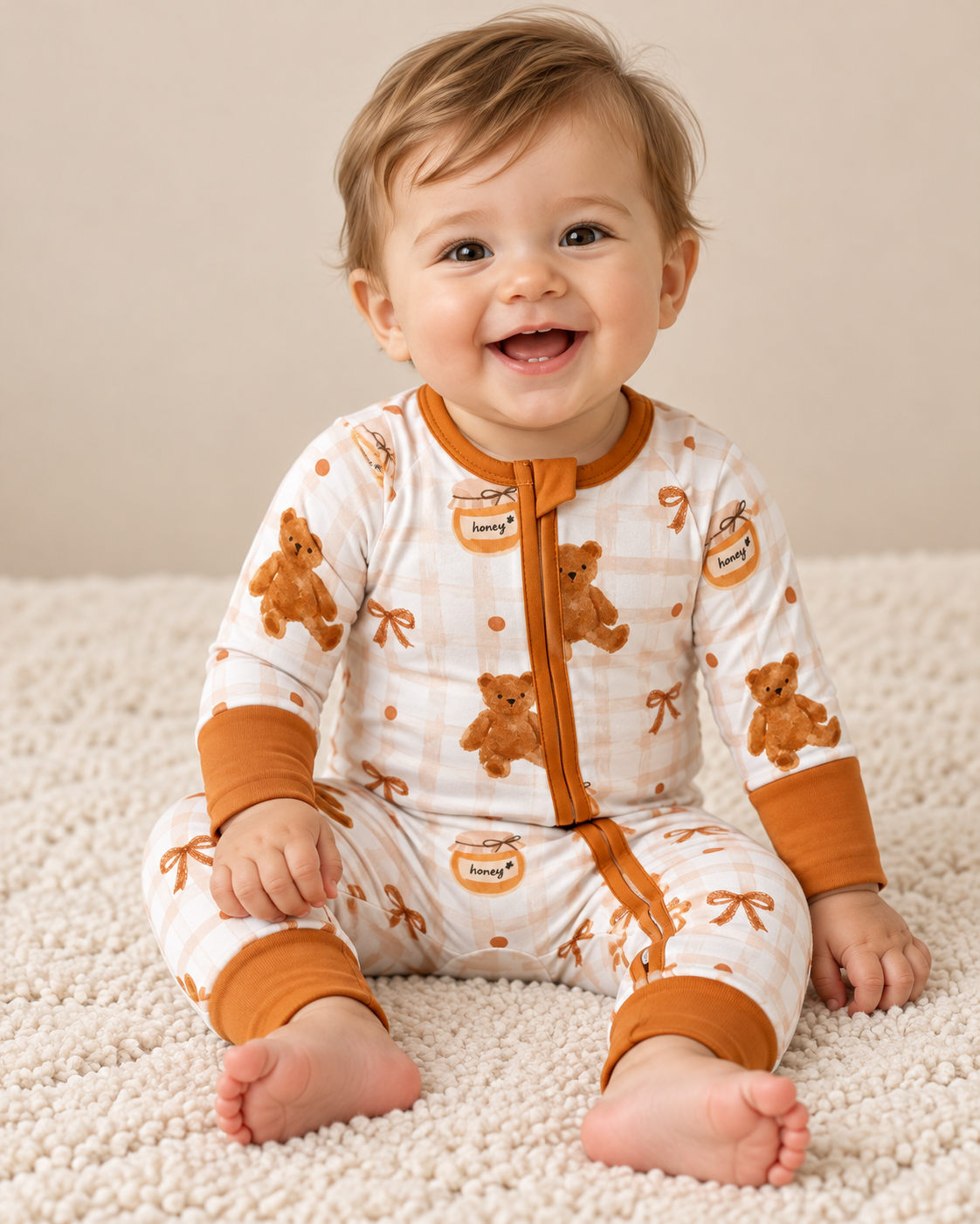 A smiling baby sits on a textured rug wearing a Honey Pot Cuddles Zippy - FULL RUN, featuring a pattern of teddy bears, honey pots, and bows.