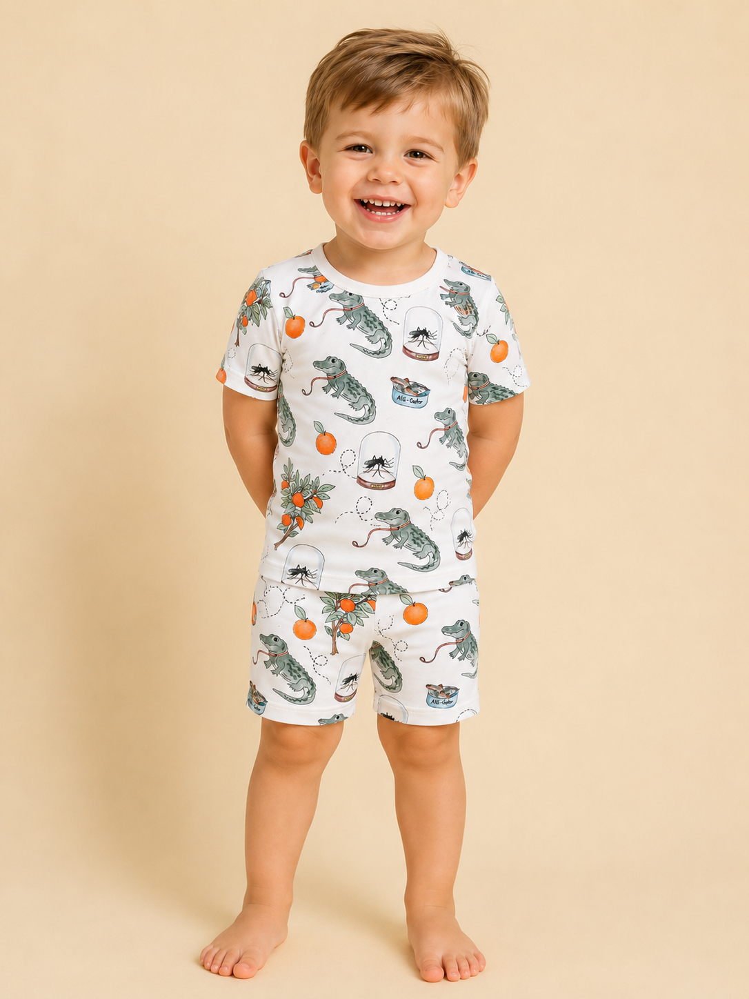 A young child smiles at the camera, barefoot and wearing the Tiny Tot Bundles Sunshine Swamp Lounge Set— a white short-sleeve pajama set with colorful alligator, orange, and flower prints on a beige background.