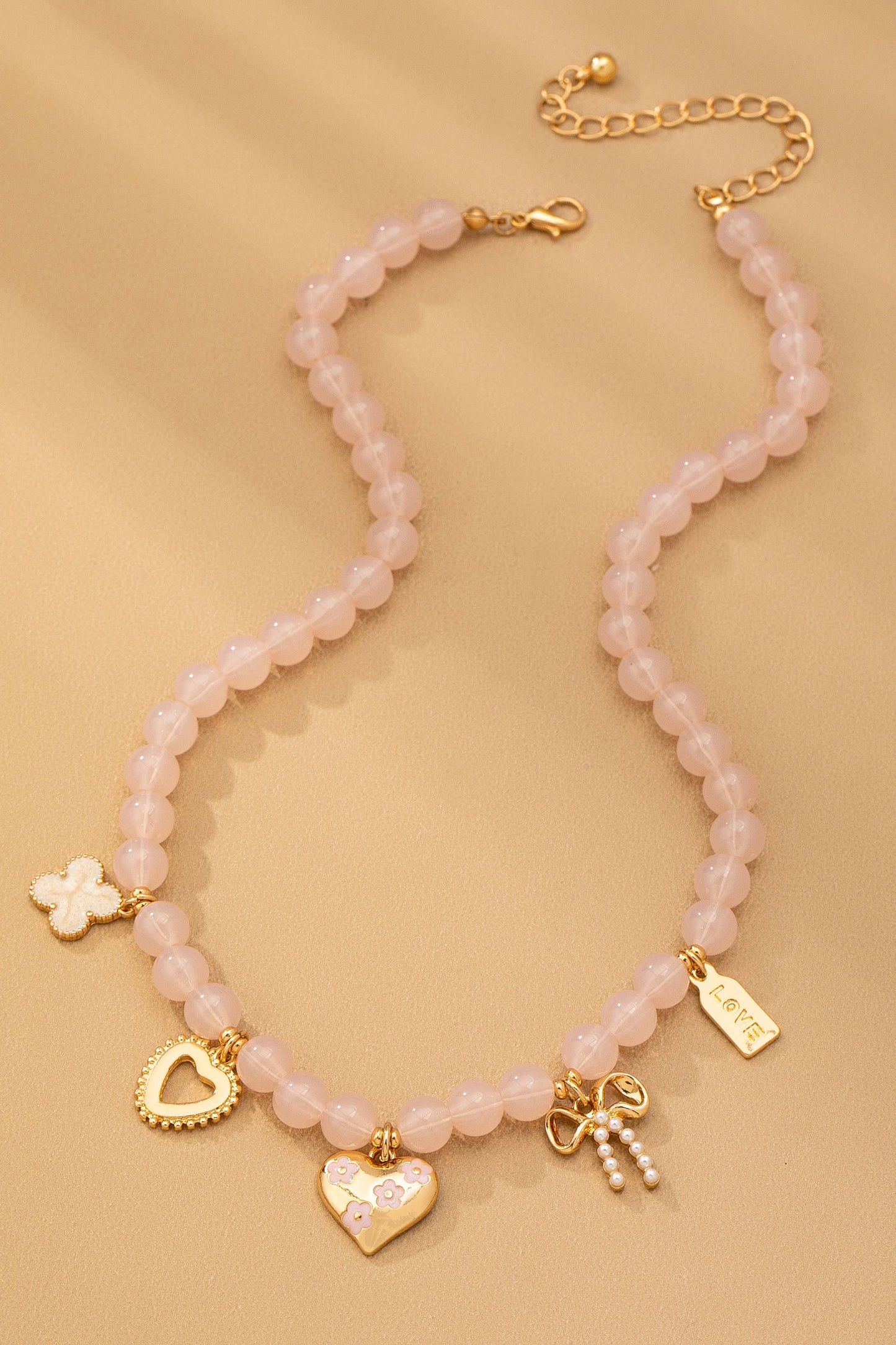 The Beaded Charm Necklace from JOSSLYN by wall to wall features pink beads and gold charms—including a heart, a bow, a four-leaf clover, a heart frame, and a rectangular LOVE tag—and is displayed on a beige surface.