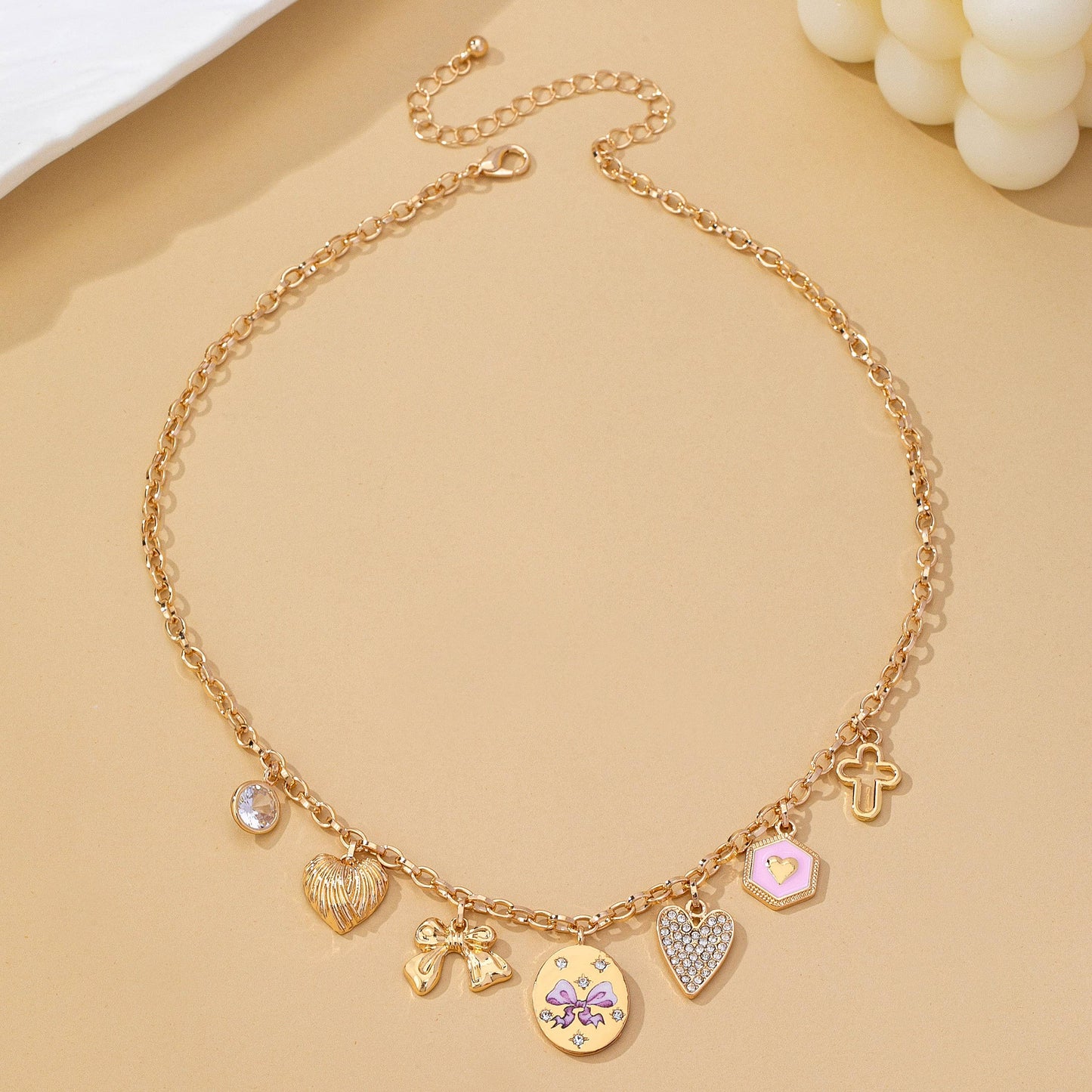 The Pink Multi-Charm Necklace by JOSSLYN by wall to wall features a gold chain adorned with various charms—including hearts, a cross, an angel, a round gem, and a disc with star designs—displayed on a beige surface.