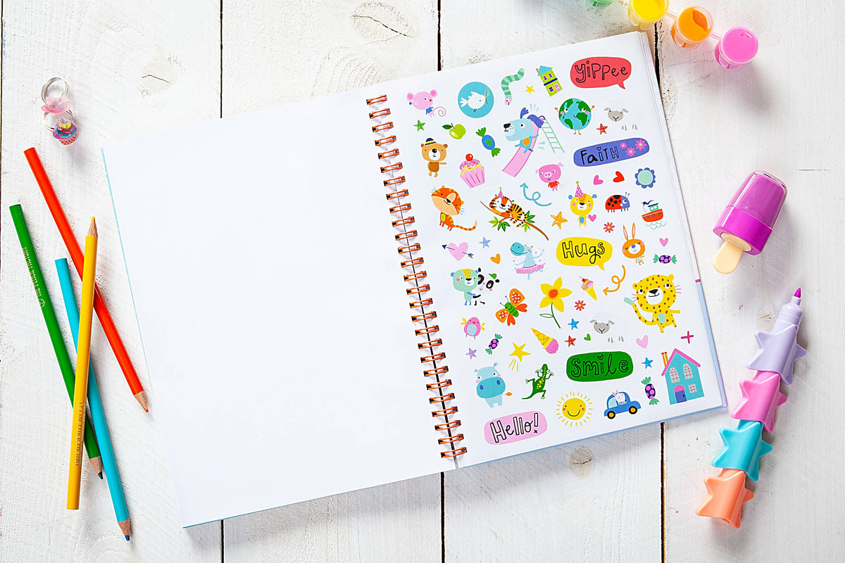 A God Made Me Journal Book by BroadStreet Publishing Group, LLC lies open on a white wooden table, its pages decorated with colorful stickers—animals, flowers, stars, and cheerful words. Nearby are colored pencils, erasers, and other stationery supplies.