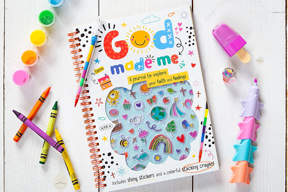 An array of vibrant supplies, including the God Made Me Journal Book by BroadStreet Publishing Group, LLC, stickers, crayons, paints, a purple pen, and a star-shaped stacking crayon, are neatly arranged on a white wooden surface.