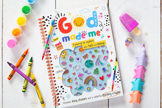 An array of vibrant supplies, including the God Made Me Journal Book by BroadStreet Publishing Group, LLC, stickers, crayons, paints, a purple pen, and a star-shaped stacking crayon, are neatly arranged on a white wooden surface.