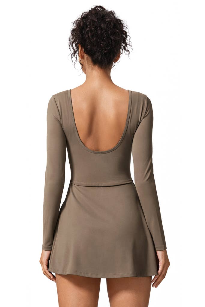A woman with curly hair in a bun stands against a white background, facing away from the camera while wearing the Color 5 Mocha Long Sleeve Athletic Dress with a low scoop back.
