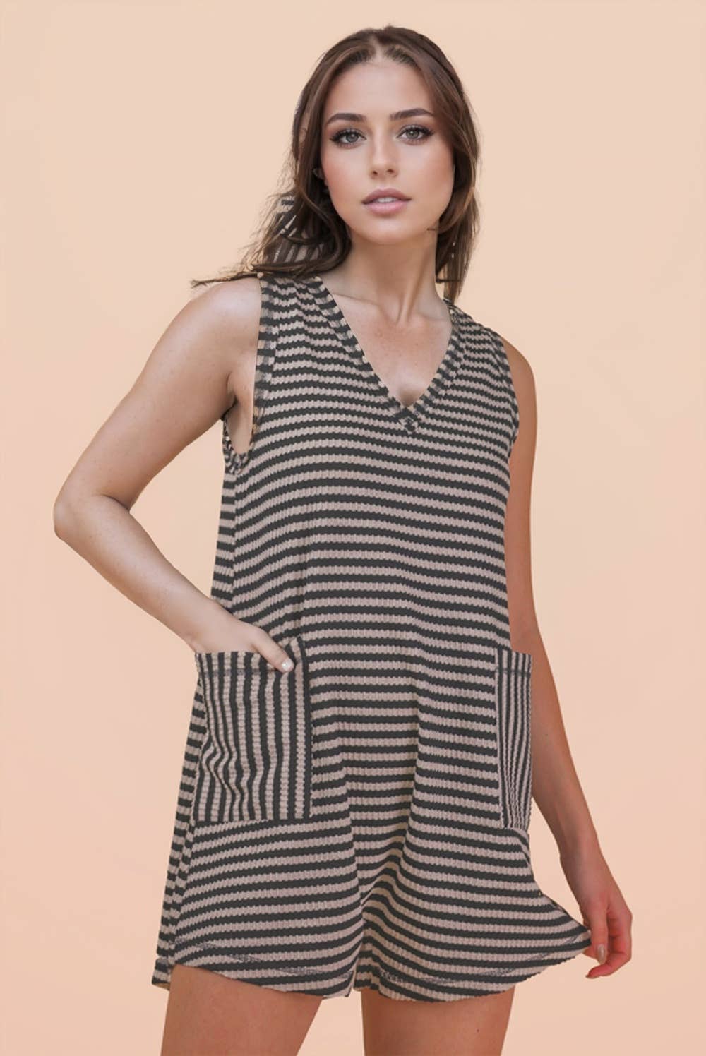 A woman wearing the VERY J Striped Knit Casual Romper, featuring a sleeveless design with black and beige stripes and large front pockets, stands against a plain peach background.