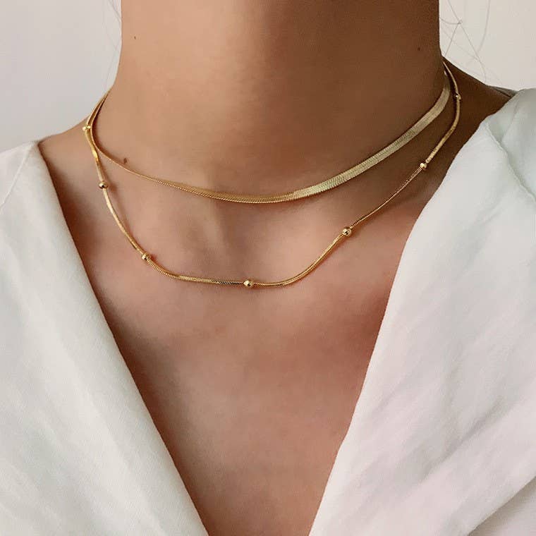 A person wears the Dainty Layered Tarnish Free Necklace by JOSSLYN by wall to wall, featuring a flat herringbone chain and a delicate chain accented with small beads.