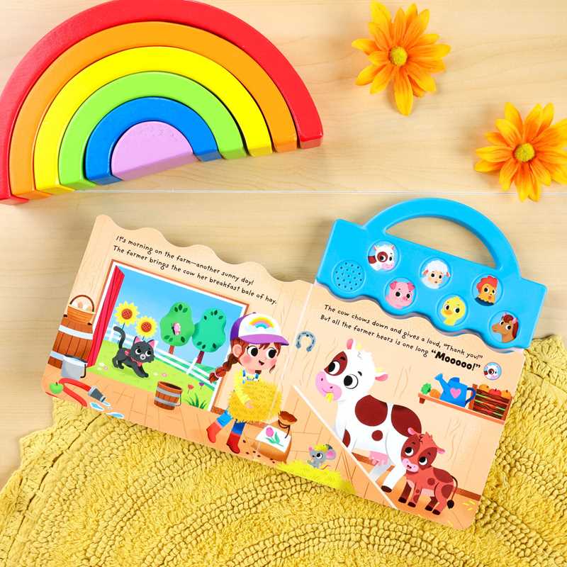The "Moo, Oink, Baa!" book by Simon & Schuster is open on a yellow mat, displaying a farm scene. Nearby are a rainbow stacking toy and two yellow flowers placed on a light wood surface.
