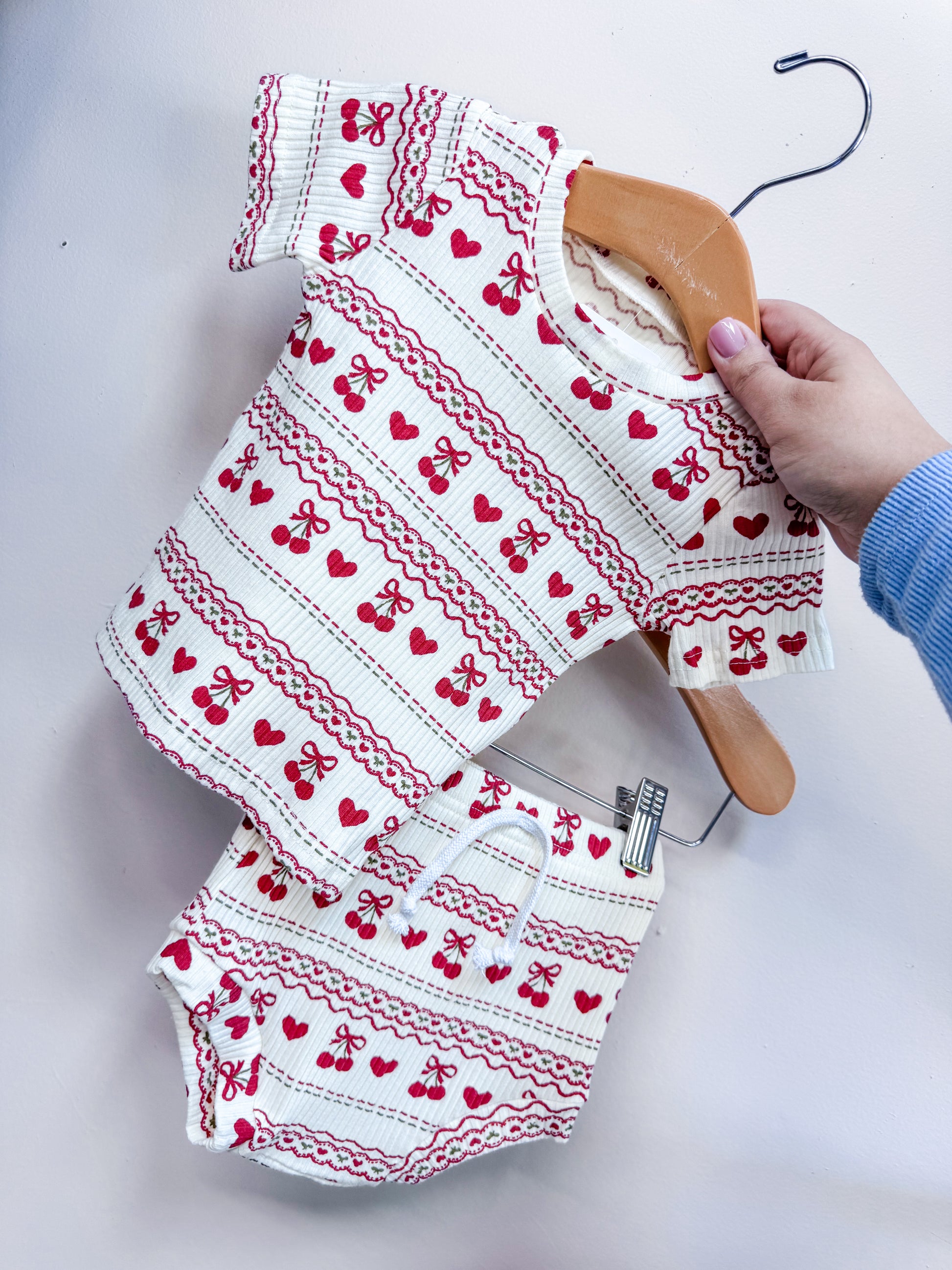 A hand holds the Cream Cherry Ribbed Set by Tiny Tot Bundles, a matching short-sleeve top and shorts featuring red cherry and bow patterns on white ribbed fabric, displayed on a hanger against a light-colored wall.