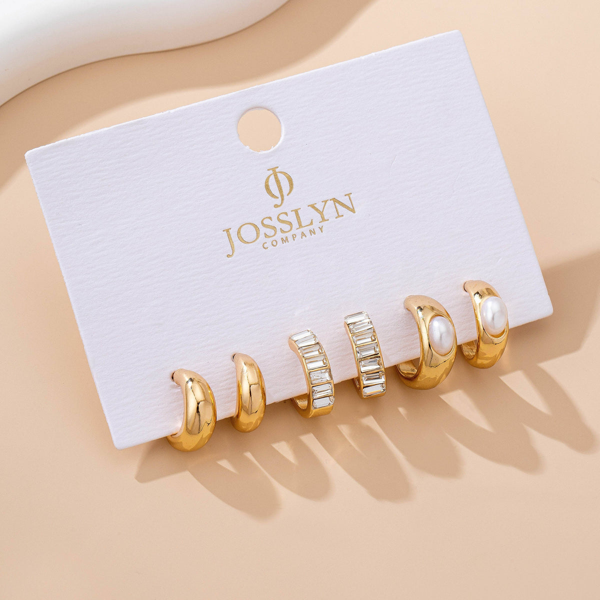 A set of five earrings from the Pearl & Baguette Hoop Trio Earring Set by JOSSLYN by wall to wall, featuring hoops, pearl, and baguette stone designs, displayed on a white company card against a beige background.