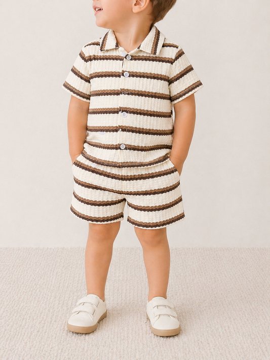 A young child stands with hands in pockets, facing slightly to the side, wearing the Tiny Tot Bundles Mocha Coast Stripe Set— a matching short-sleeve, button-up shirt and shorts with cream and brown stripes—paired with cream-colored shoes.
