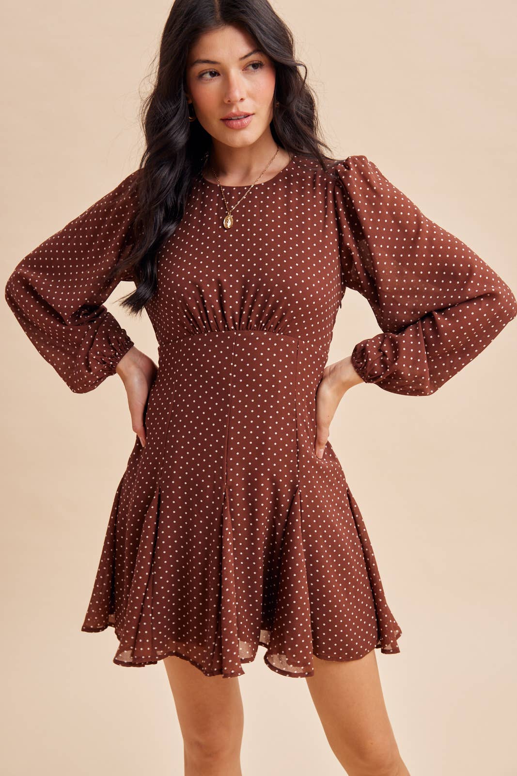 A woman wears an In Loom Polka Dot Flared Mini Dress, featuring a brown long-sleeved design with a white polka dot pattern, a fitted bodice, and a flared skirt.