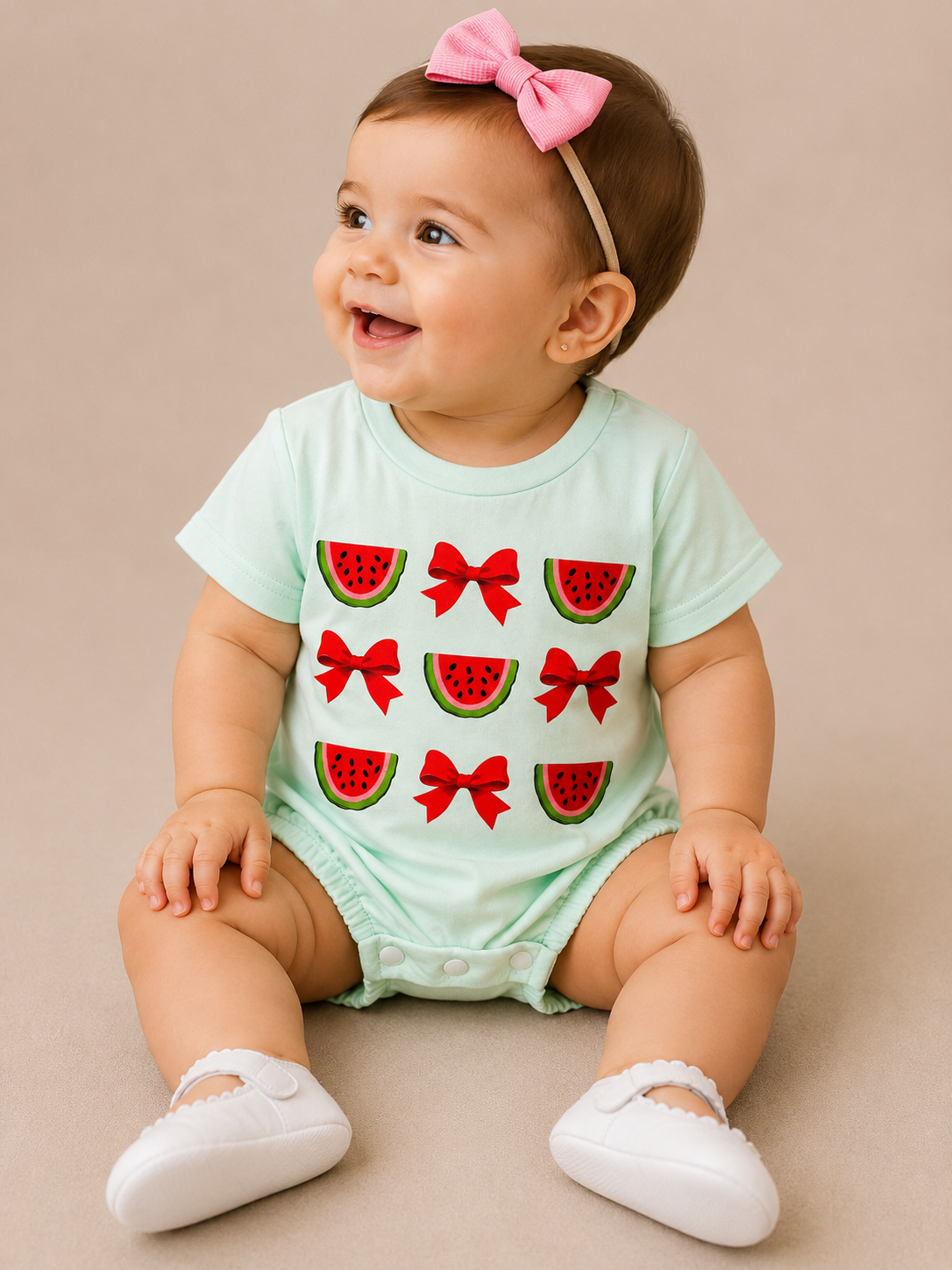 A smiling baby in the Tiny Tot Bundles Watermelon Bow Bubble, with red watermelon and bow designs, a pink headband, and white shoes, sits on a beige background.