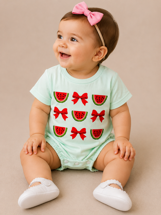 A smiling baby in the Tiny Tot Bundles Watermelon Bow Bubble, with red watermelon and bow designs, a pink headband, and white shoes, sits on a beige background.