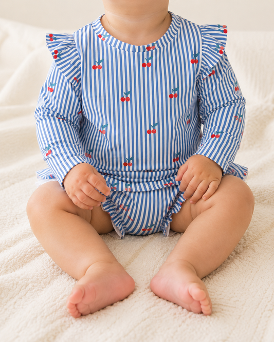 A baby, face not visible, sits on a light-colored blanket wearing the Tiny Tot Bundles Cherry Stripe Swim Romper, a blue and white striped outfit with red cherry prints and ruffled sleeves.