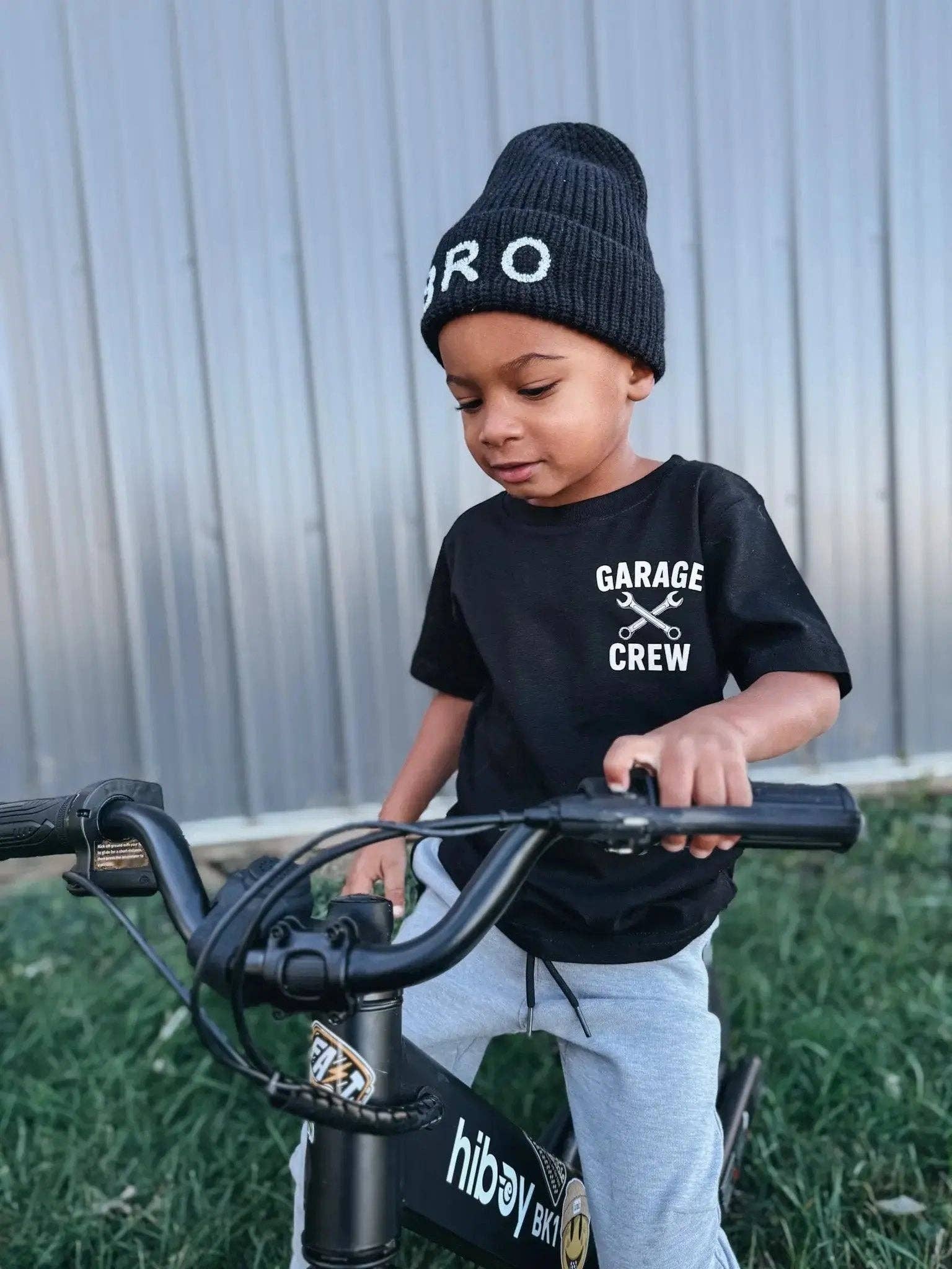 A young child wearing a black beanie labeled "BRO" and a Raising Brave My Dad's Garage Toddler Tee sits on a black Hiboy bike, looking down while outdoors on grass with a metal wall in the background.