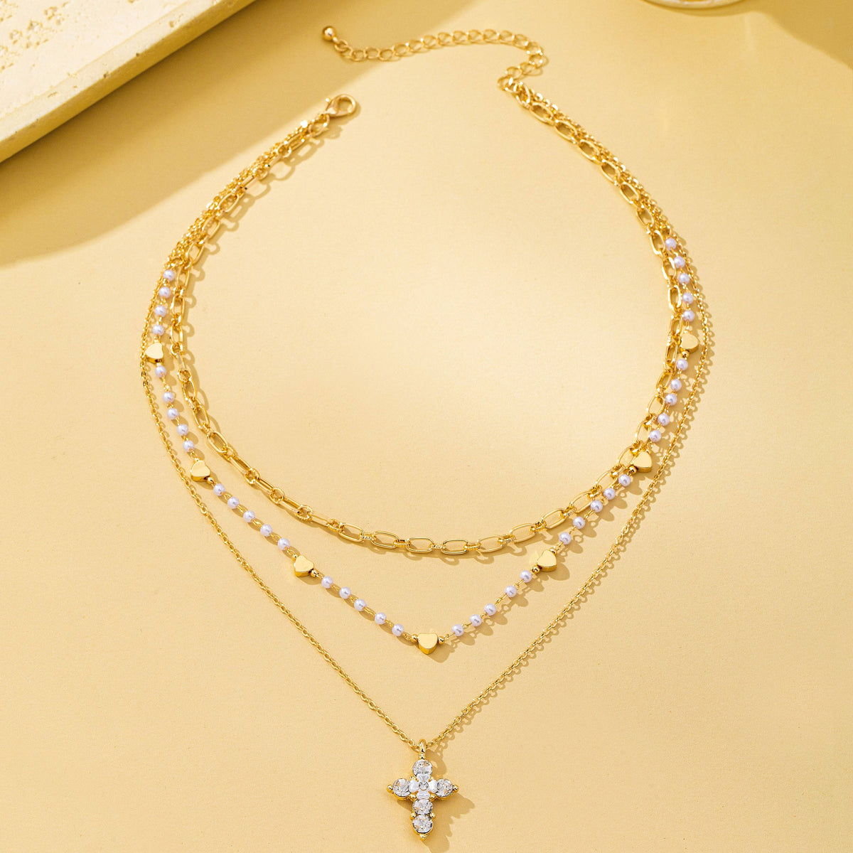 JOSSLYN by wall to wall’s Multi-Strand Pearl & Crystal Cross Necklace features three gold chains: one adorned with small gold hearts, another with pearls and hearts, and the shortest chain showcasing a cross pendant embellished with sparkling white stones, all beautifully displayed on a light beige background.