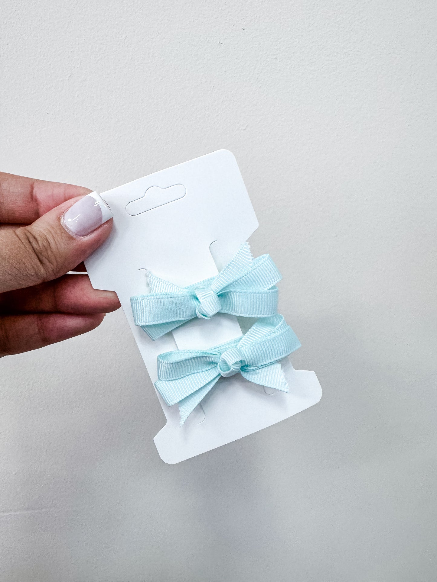 A hand with manicured nails holds a white card displaying the Duo Piggy Clip Pack by Tiny Tot Bundles—two light blue ribbon bows—against a plain light background.