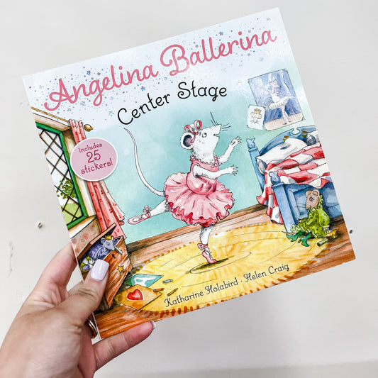 A hand holds the Simon & Schuster Angelina Ballerina Center Stage Book, showing an illustrated mouse in a pink tutu dancing in a colorful bedroom with a frog on the bed and a window in the background.