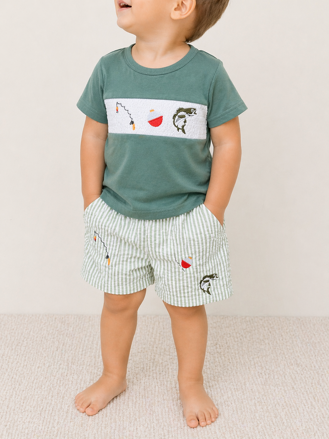 A young child stands barefoot on a light carpet, wearing Tiny Tot Bundles' Little Angler Short Set featuring a green tee with fishing embroidery and coordinating green-and-white striped shorts adorned with colorful lure accents.