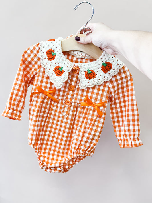 A hand holds the Tiny Tot Bundles Crochet Pumpkin Gingham Romper, featuring orange and white gingham, orange bows, and a white crocheted collar adorned with small orange pumpkins.