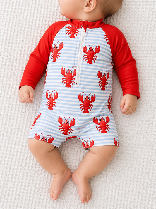 A baby lies on a cream textured blanket wearing the Tiny Tot Bundles Red Claw Bay Swimsuit, featuring red sleeves, cartoon lobsters, and a front zipper. The baby's face is not visible.