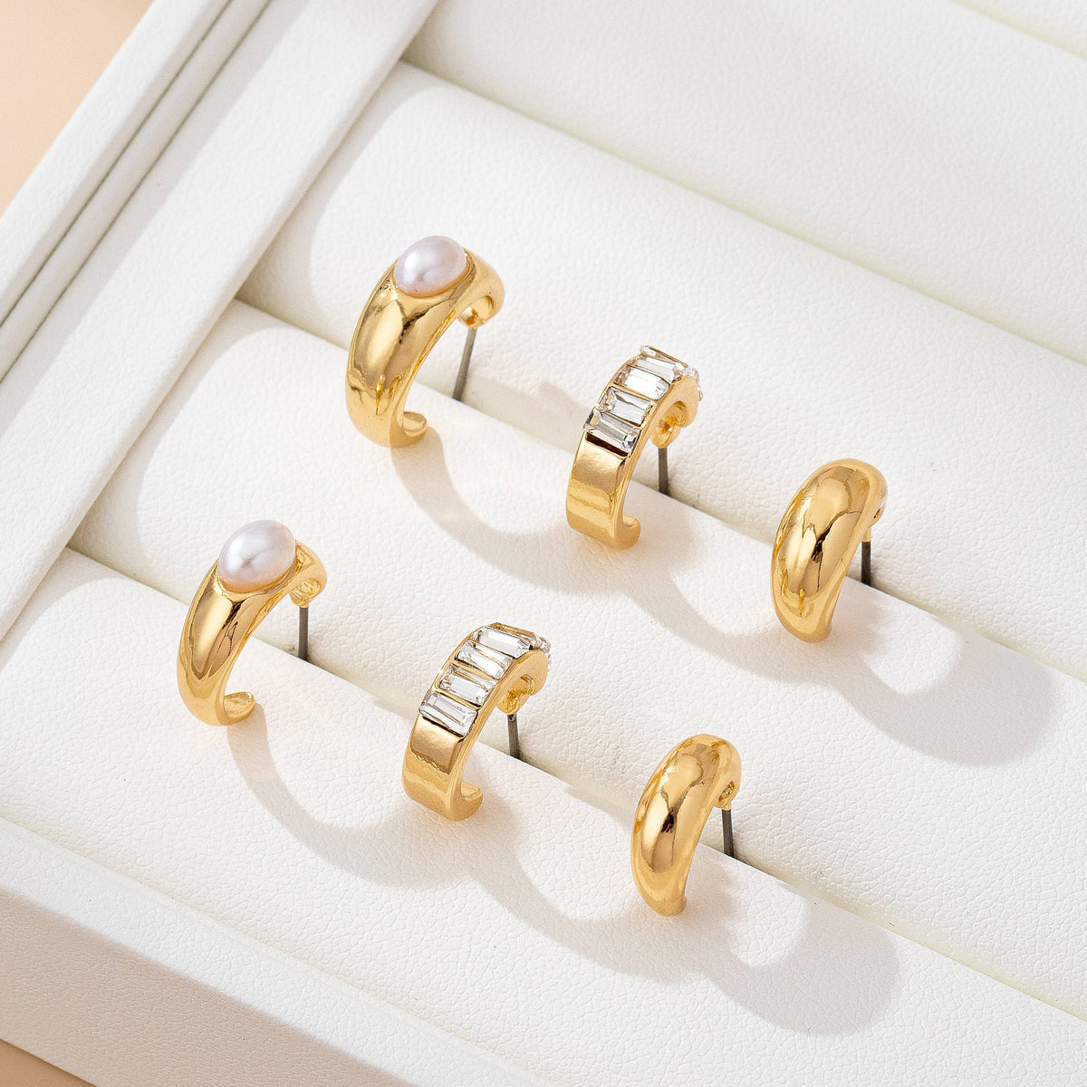 Three pairs of gold hoop earrings from the JOSSLYN by wall to wall Pearl & Baguette Hoop Trio Earring Set, featuring pearls and rectangular gemstones, are displayed in a white jewelry tray.