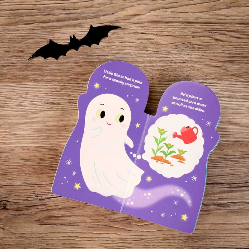 The “Little Ghost by Maggie Fischer” book from Simon & Schuster, shaped like a ghost, is open on a wooden table displaying a ghost illustration and a thought bubble featuring plants and a watering can, with a black bat decoration placed nearby.