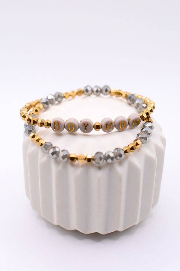 The Boy Mom Bracelet Set by Two and Crew - Faith Based Jewelry & Gifts is displayed on a white textured stand, featuring one bracelet with white letter beads spelling “BOY MOM” and another adorned with gold and silver beads.