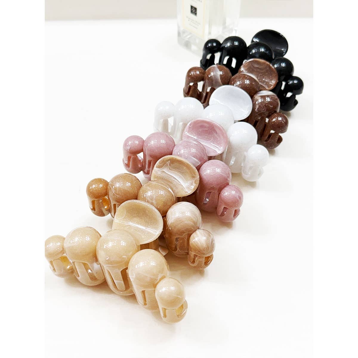 Several Love and Repeat Large Marbled Hair Claws in an assortment of colors—beige, brown, pink, white, and black—are neatly arranged in rows on a white surface. A glass perfume bottle is partially visible in the background.