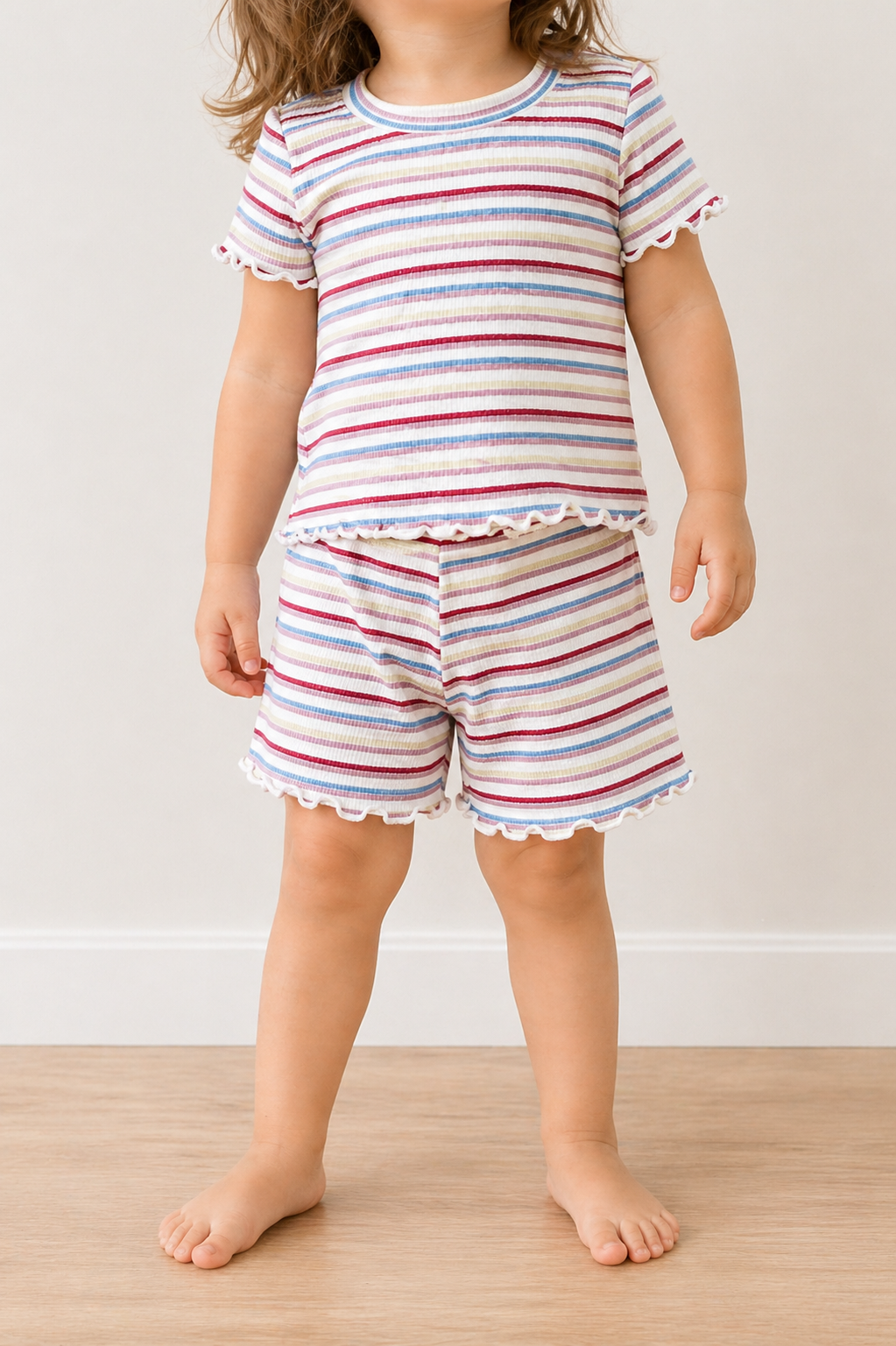 A young child wears the Tiny Tot Bundles Sweet Stripe Snuggle Set, featuring a matching short-sleeved top and shorts with pastel stripes and ruffled edges, standing barefoot on a wooden floor against a plain light background.